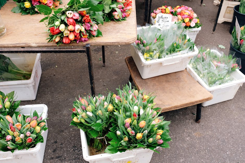 A variety of fresh tulip bouquets in shades of pink, yellow, white, and red with green foliage, arranged in white plastic crates and placed on and around small wooden tables. The tulips have smooth, c
