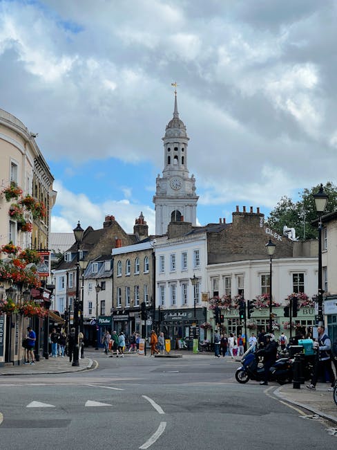The image depicts a lively street scene with a historic white clock tower with a gold weather vane at the top, set against a partly cloudy sky. Surrounding the tower are multi-storey buildings with tr