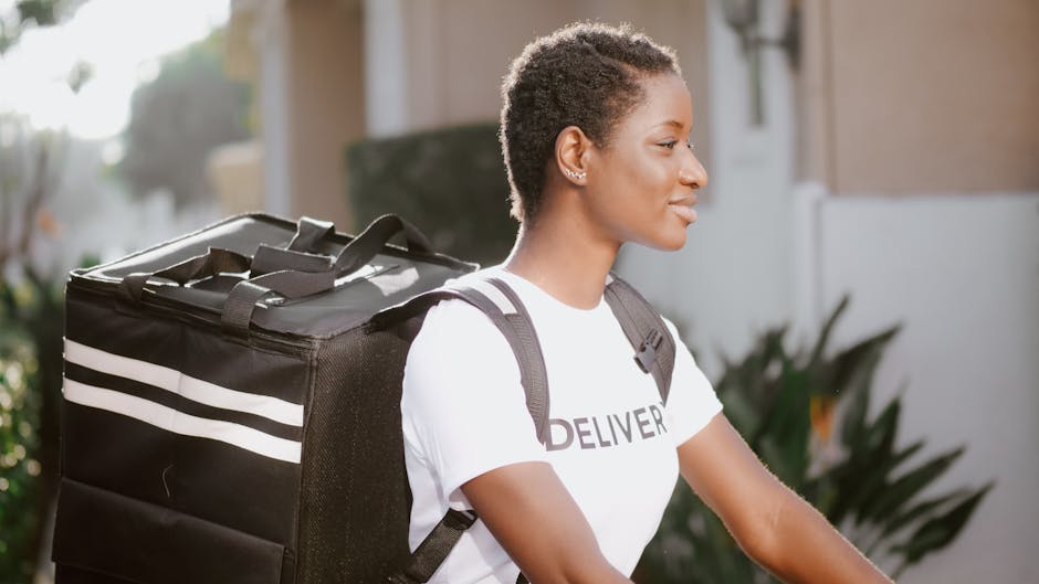 A young woman with short, natural hair walks outdoors in natural daylight, wearing a white T-shirt with the word 'DELIVER' printed on it and carrying a large black delivery bag with white stripes on h