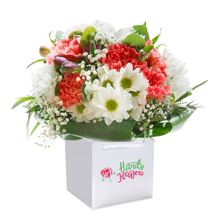 Standing perfectly upright in a sharp white gift bag designed for elegance and convenience, this expertly curated bouquet channels the vivacious spirit of Shepherd's Bush. Plush coral and peach carnations, instantly recognizable for their layered, ruffled petals, blend seamlessly with crisp, daisy-like white chrysanthemums featuring vivid lime-green centers. Delicate sprigs of baby's breath weave through the bouquet, casting an ethereal softness and echoing the airy light filtering through local parks. A few tight, purple-tinged buds hint at freshly emerging blooms, adding intrigue and unseen promise. Deep, glossy green leaves enclose the arrangement in a natural, verdant border, providing both structure and contrast reminiscent of the garden squares near Uxbridge Road. The bouquet's perfectly rounded form, coupled with the substantial foliage, showcases a balanced interplay between lush fullness and delicate lightness. Presented against a spotlessly pale background, the bouquet's colours and textures leap forward, inviting admiration for both its freshness and craft. Ready for same-day delivery across Shepherd's Bush, this joyous arrangement is ideal for birthdays, anniversaries, or any occasion when a heartwarming, thoughtfully presented floral gift is called for-each petal and leaf a testament to the skilled hands of your local florist.